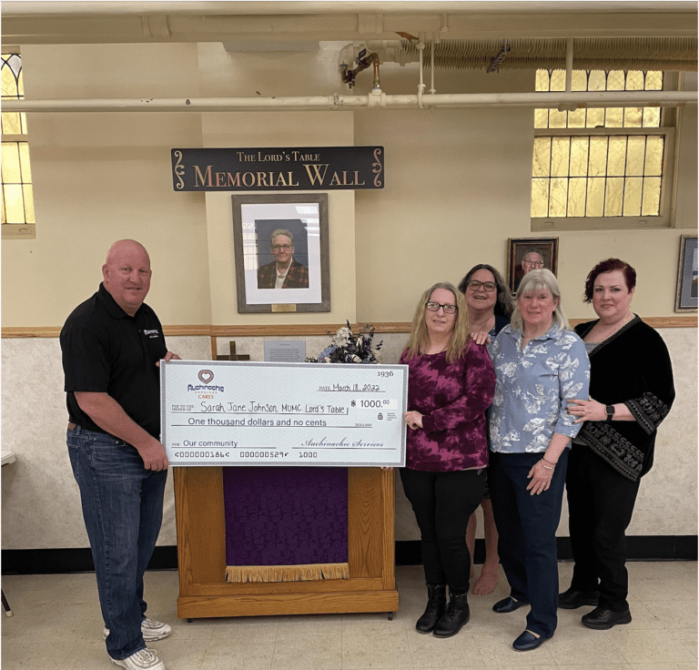 Five people stand indoors, proudly holding a large ceremonial check for $1,000 in front of a memorial wall, embodying the spirit of generosity that defines our 'About Us' story.