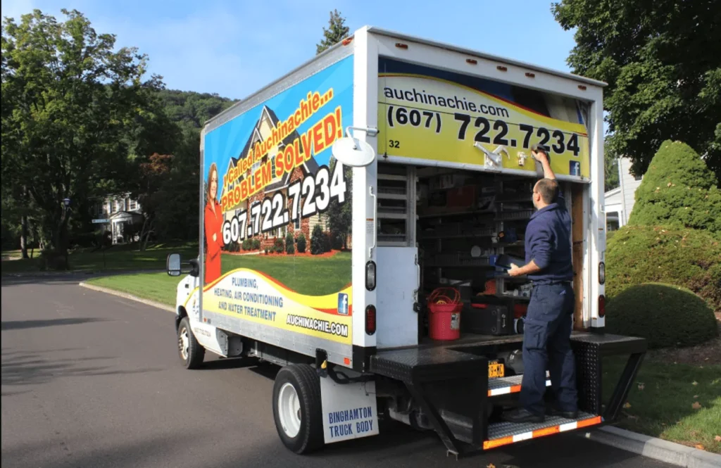 A worker stands at the back of a plumbing service truck with its doors open, organizing tools and equipment on a residential street.