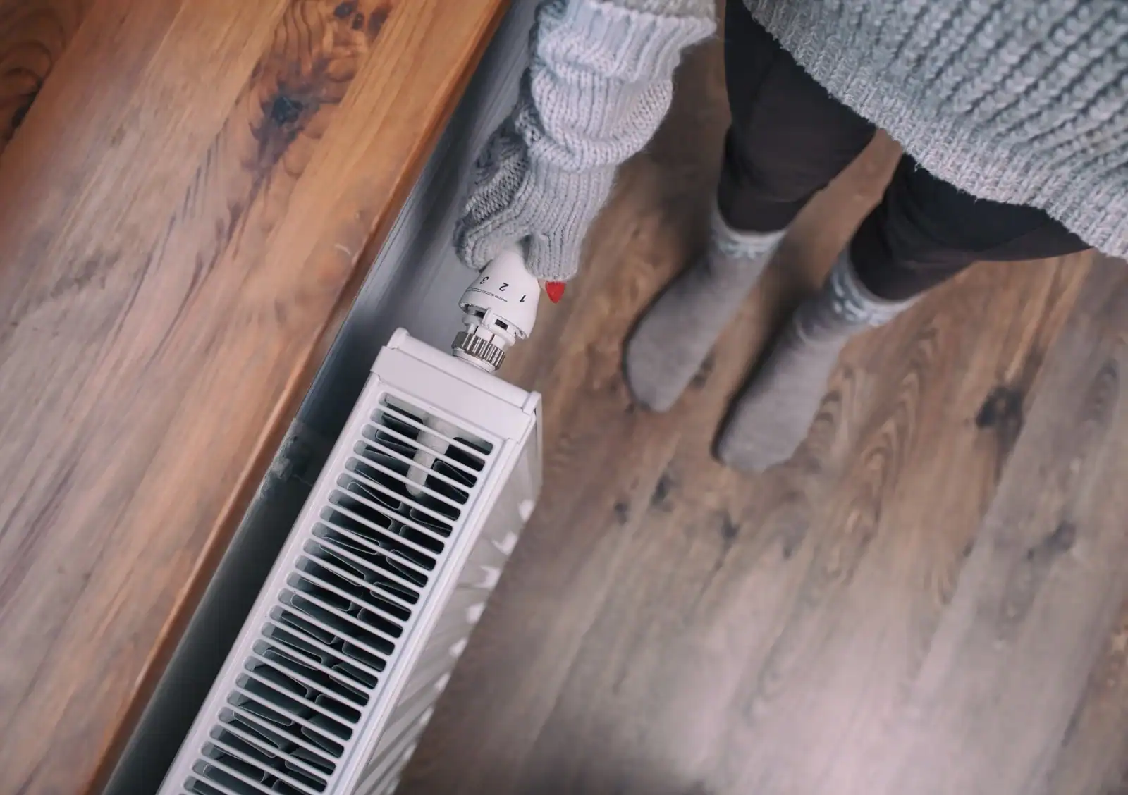 A person wearing a gray sweater and gloves adjusts a thermostat knob on a white radiator. They are standing on a wooden floor.