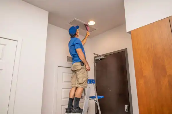 A person wearing a blue shirt and khaki shorts stands on a ladder, changing an air filter in a ceiling vent inside a room.