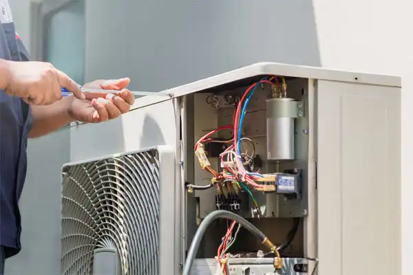 A technician uses a screwdriver to repair an open air conditioning unit with visible colorful wires and components.