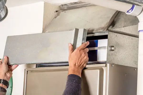 Person removing a metal panel from a furnace, exposing the interior components, in a maintenance or repair scenario.