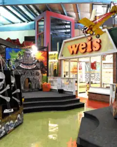 Indoor play area featuring a castle-themed structure on the left, a Weis storefront on the right, and a yellow toy airplane suspended from the ceiling—brought to you with support from Auchinachie Cares.