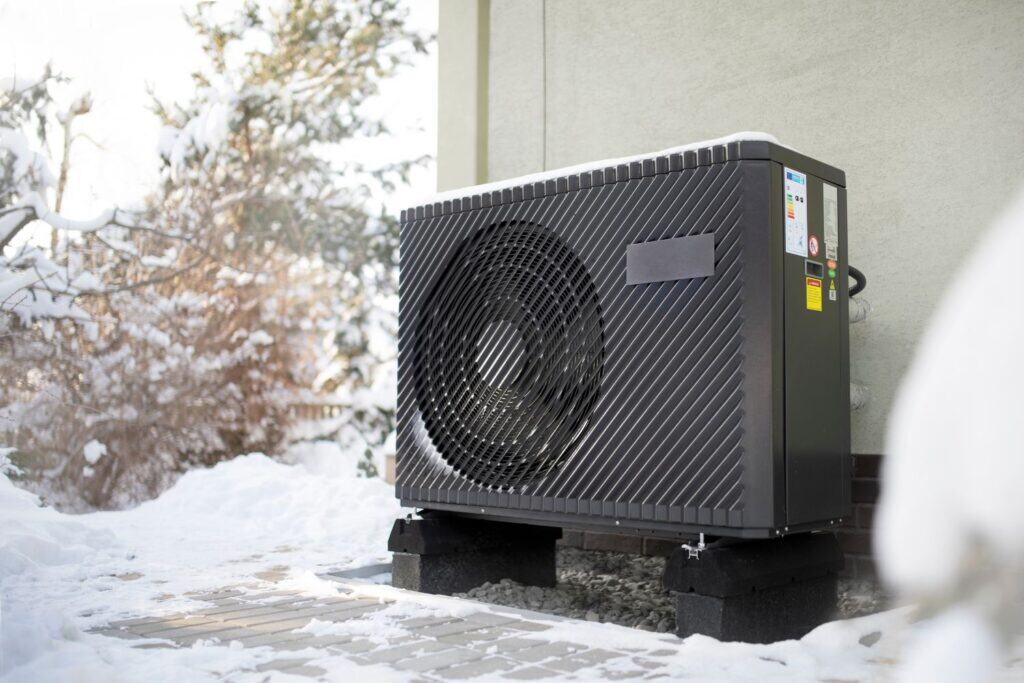 A heat pump unit installed outside a building in a snowy area, with snow covering the ground and trees in the background.