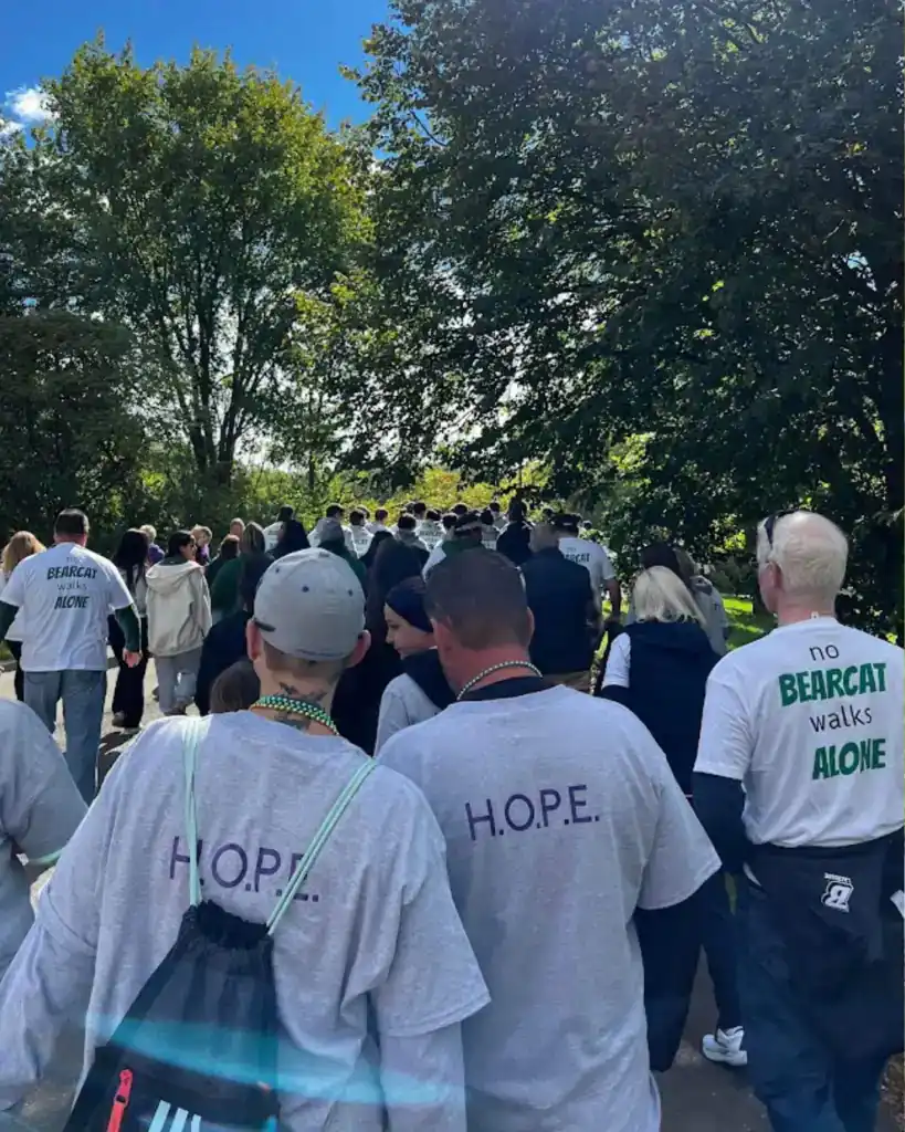 A group of people walk together outdoors, some wearing shirts with "H.O.P.E.," "No Bearcat walks alone," and “Auchinachie Cares,” surrounded by trees on a sunny day.
