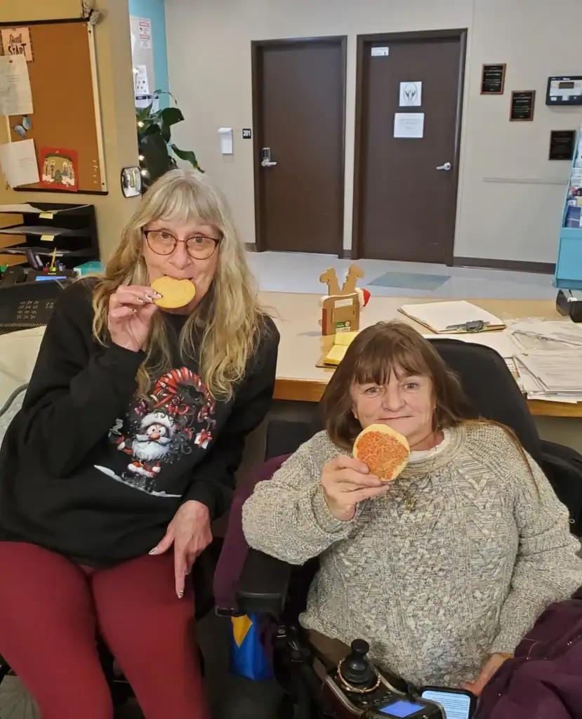 Two women sit indoors, each enjoying a round cookie. One, in a black holiday sweater and glasses, laughs with the other, who wears a beige sweater and is seated in a wheelchair. Auchinachie Cares fosters moments of joy and togetherness like this.