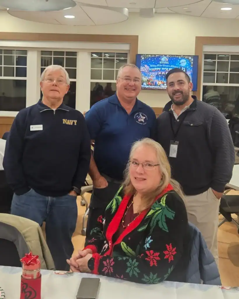 Four adults pose for a group photo indoors; three men stand behind a seated woman at a table decorated for the holidays, showcasing the spirit of Auchinachie Cares.