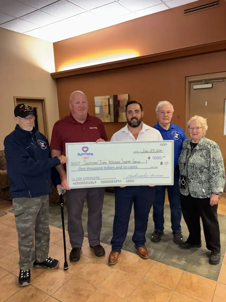Five adults stand indoors holding a large ceremonial check for $1,000 made out to the Veterans Support Group, presented by Auchinachie Cares.