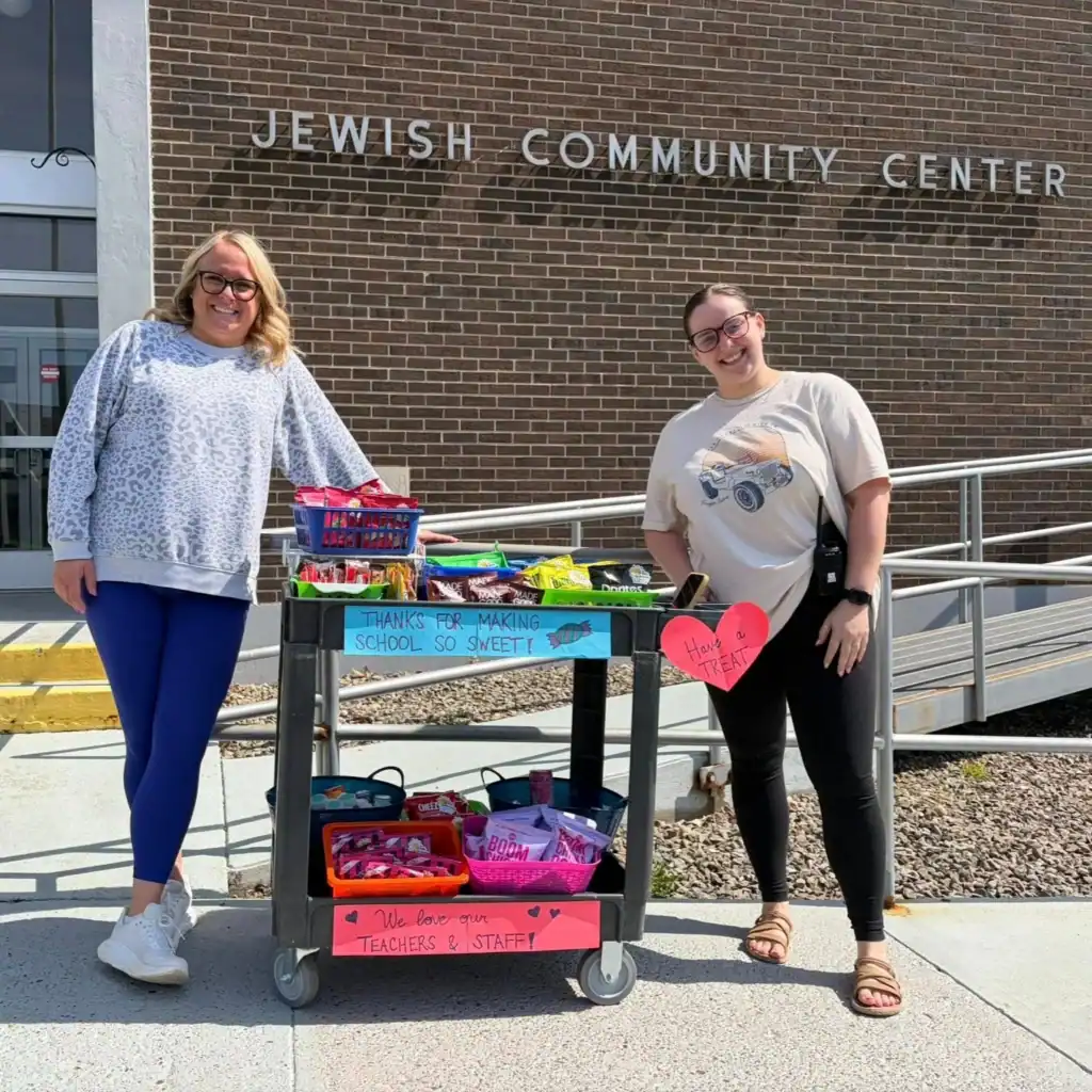Two women stand beside an Auchinachie Cares snack cart with thank-you signs in front of a Jewish Community Center building.