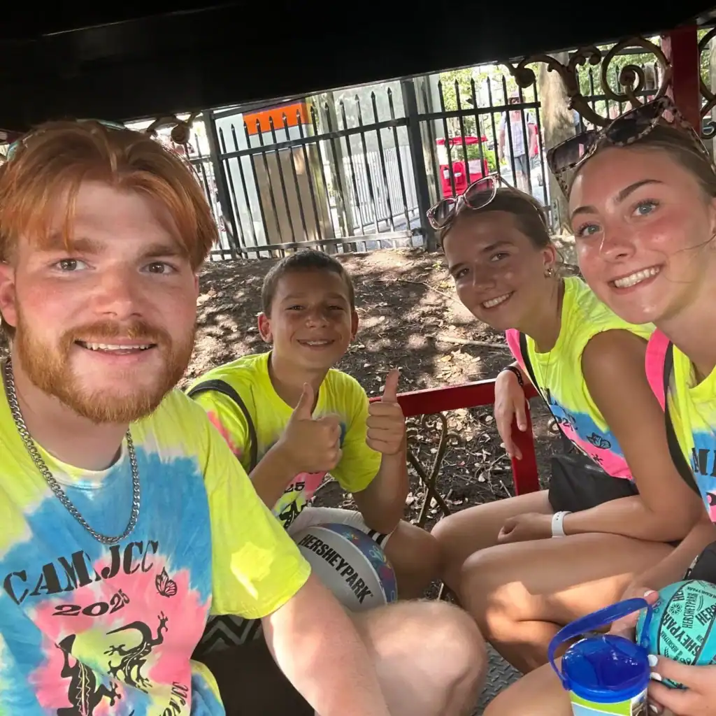 Four young people in bright tie-dye shirts sit together in a shaded area, smiling at the camera. One boy gives a thumbs up, showing the fun spirit of Auchinachie Cares. A fence and trees are visible in the background.