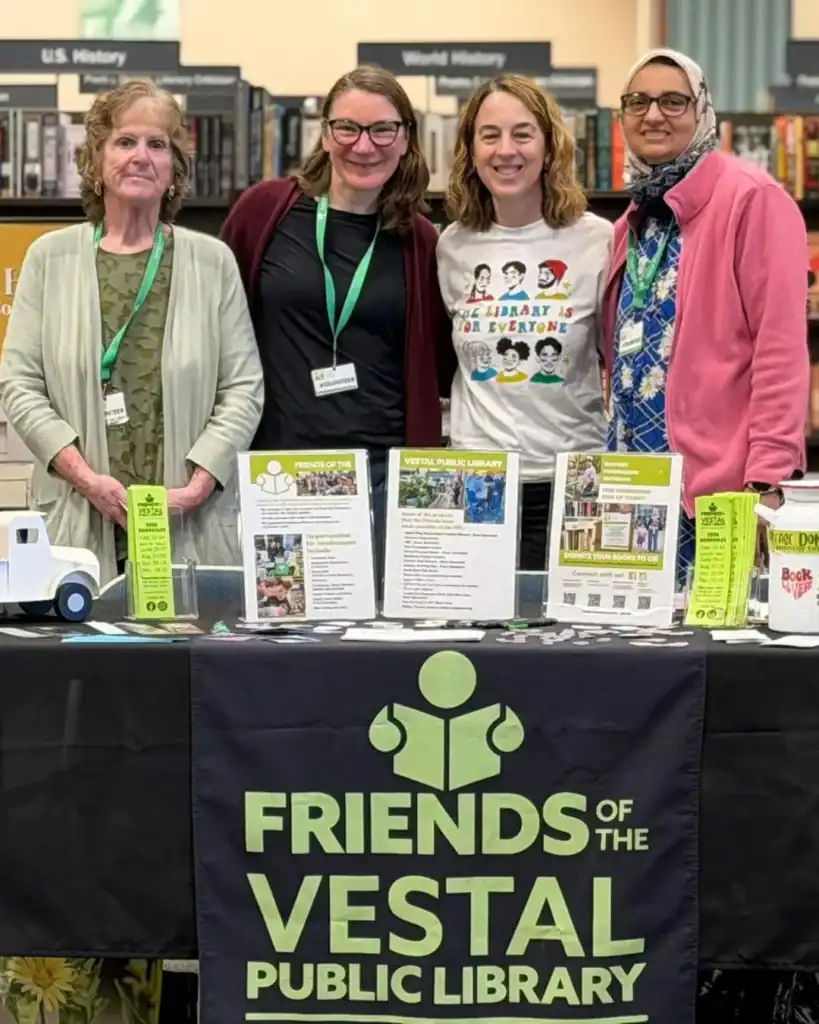 Four Auchinachie Cares volunteers stand behind a table at a Friends of the Vestal Public Library event, displaying information pamphlets and a donation jar.