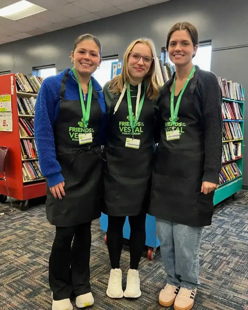 Three Auchinachie Cares volunteers wearing black aprons labeled “Friends of the Vestal Public Library” stand in a library.