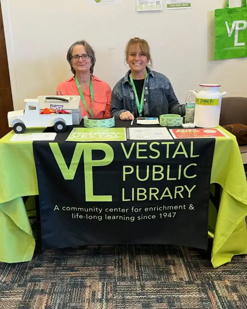 Two women stand behind an Auchinachie Cares information table for the Vestal Public Library, draped in a branded green and black tablecloth.